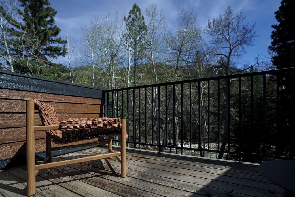 Una terraza de madera con un banco de mimbre, baranda de metal negro y vistas a árboles y colinas.