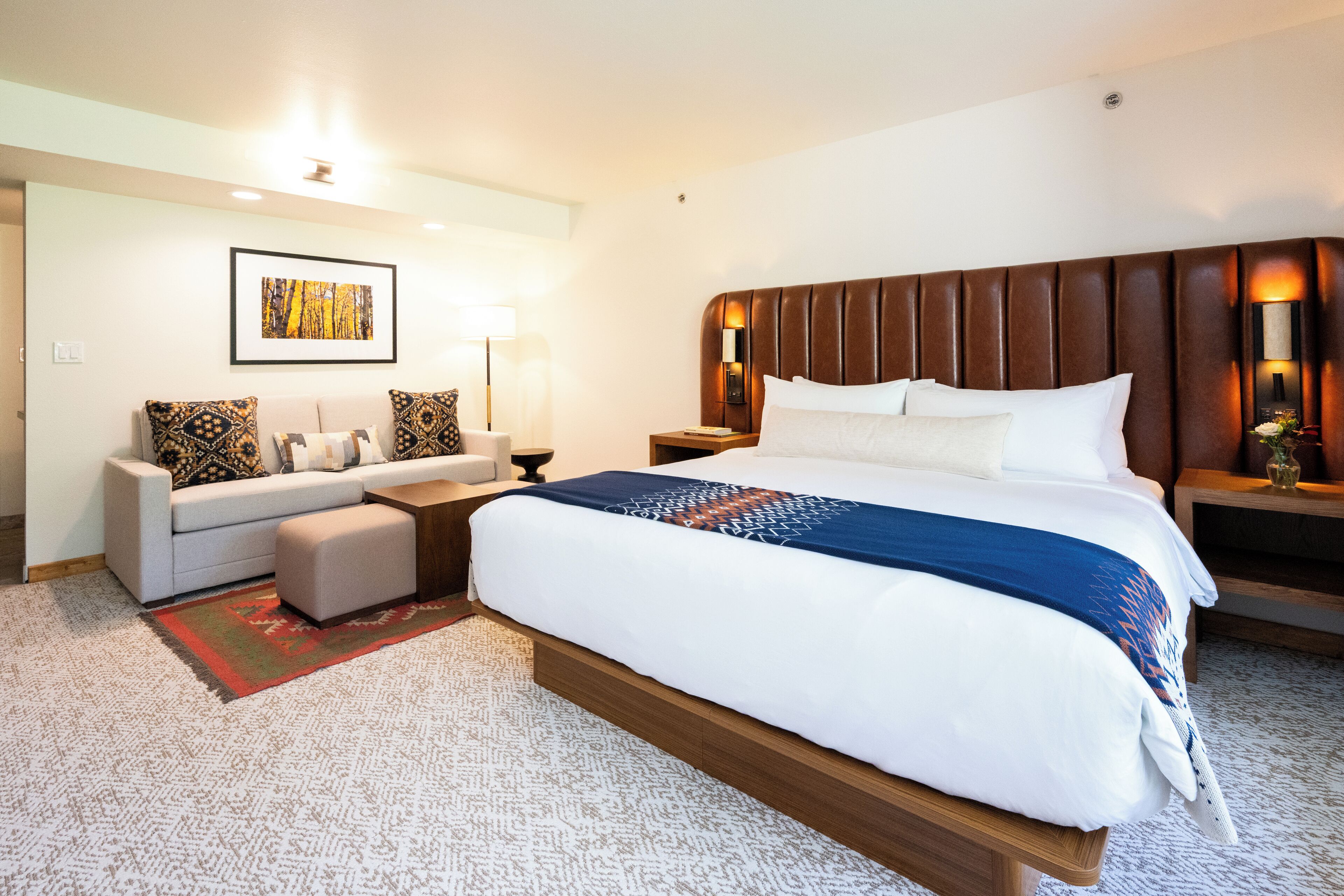 A hotel room with a large bed, a sofa, a coffee table, and a side table with a lamp.