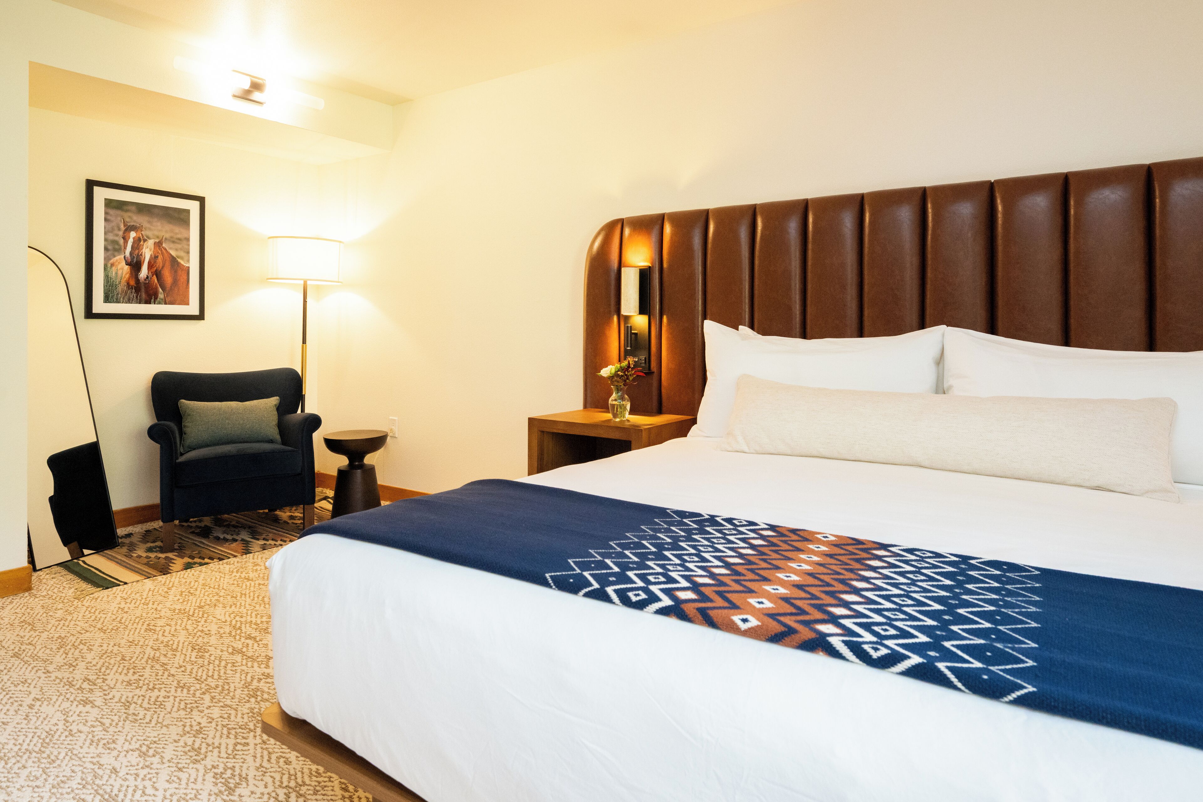 A hotel room with a large bed, a headboard, a nightstand with a lamp, a chair, and a framed picture on the wall.