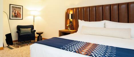 A hotel room with a large bed, a headboard, a nightstand with a lamp, a chair, and a framed picture on the wall.