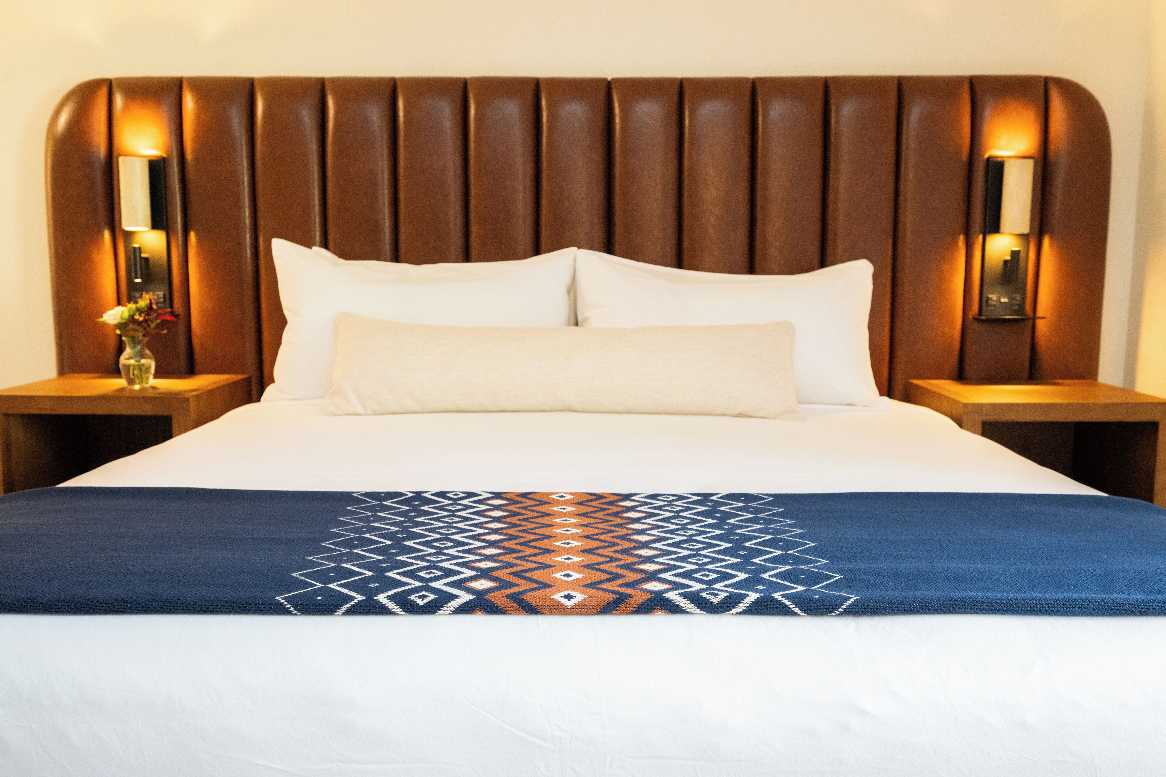 A hotel room with a large bed, a headboard with vertical slats, two bedside tables with lamps, and a decorative throw blanket.