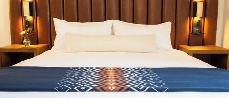 A hotel room with a large bed, a headboard with vertical slats, two bedside tables with lamps, and a decorative throw blanket.