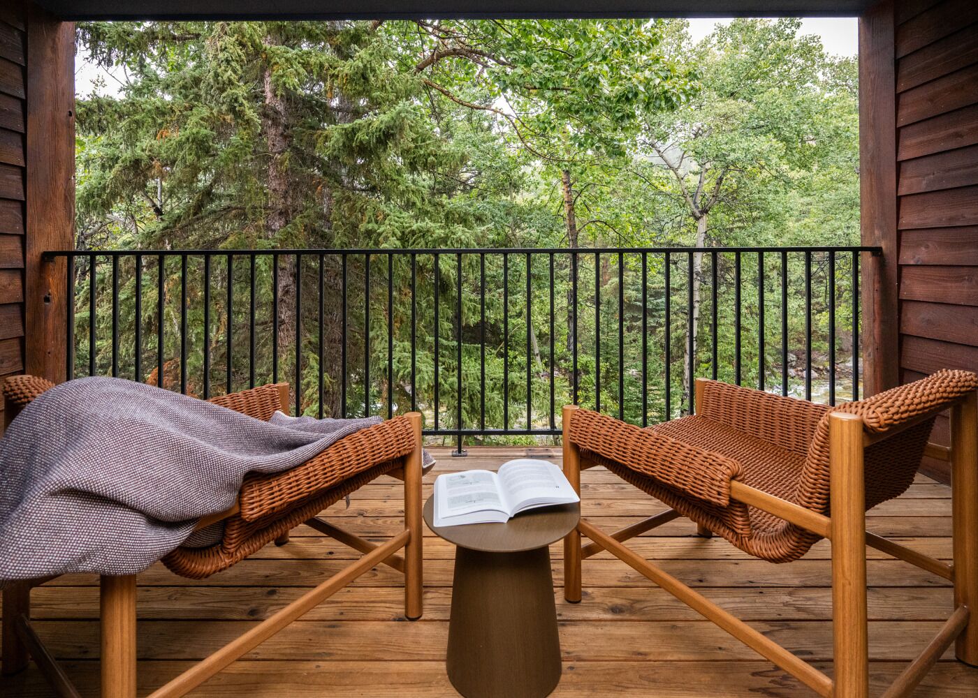 A wooden deck with two wicker chairs and a table, overlooking a forested area.