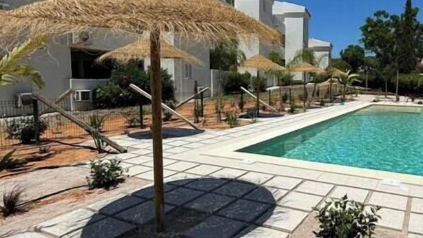 A poolside area with a clear blue pool, surrounded by white buildings and thatched umbrellas.