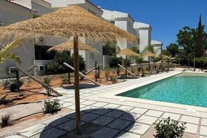 A poolside area with a clear blue pool, surrounded by white buildings and thatched umbrellas.