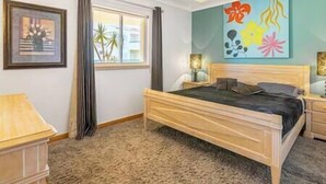 A bedroom with a wooden bed, a nightstand, a lamp, a window with curtains, and a colorful abstract painting on the wall.