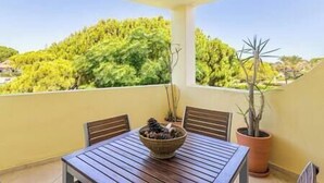 A balcony with a wooden table, chairs, a potted plant, and a view of green trees.