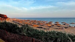 A beach with numerous umbrellas and sun loungers, a clear sky, and a rocky coastline.