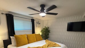 Una cama bien hecha con ropa de cama blanca y amarilla, un ventilador de techo, una mesita de noche con lámpara y un televisor montado en la pared.