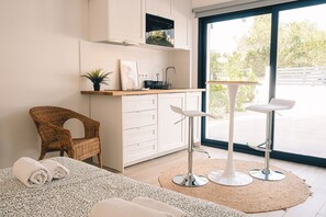 Une cuisine moderne avec des meubles blancs, un four à micro-ondes et une petite plante. On y trouve une chaise en osier, un lit avec des serviettes pliées et deux tabourets de bar blancs.
