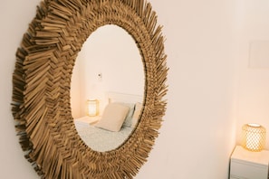 A room with a large woven mirror, a bed with patterned bedding, and two wall-mounted lamps.
