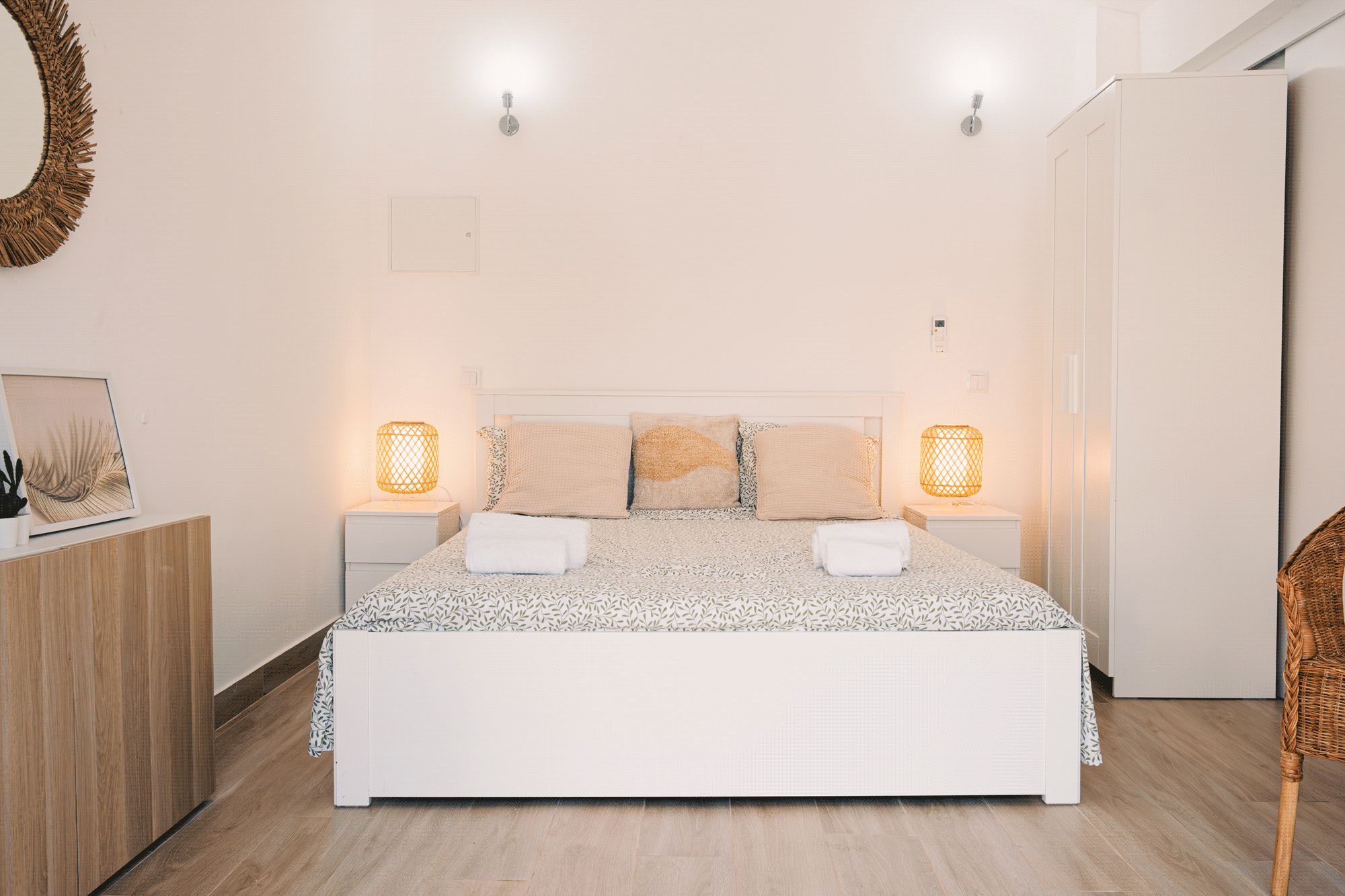 Een keurig opgemaakt slaapkamer met een bed, bedlampjes, een ingelijst beeld en een rieten stoel.
