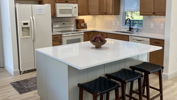 A modern kitchen with wooden cabinets, a white island with bar stools, a refrigerator, a microwave, and a window with a view. - Renovated! Between Two Squares with Golf Cart ! (The Villages)