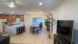 A modern living room with a flat-screen TV, a dining area with a table and chairs, a kitchen with wooden cabinets, and a ceiling fan. - Renovated! Between Two Squares with Golf Cart ! (The Villages)