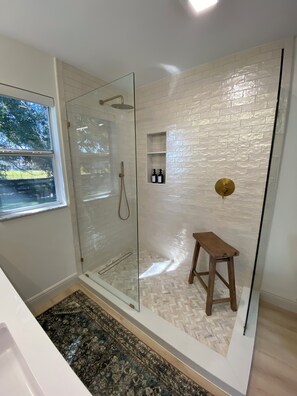A modern bathroom with a glass-enclosed shower, a wooden stool, and a small shelf with toiletries. - Renovated! Between Two Squares with Golf Cart ! (The Villages)