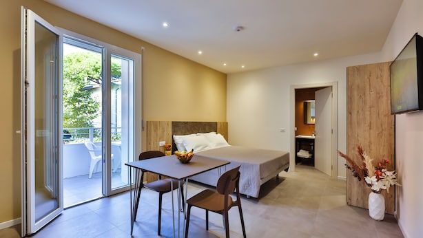 A modern hotel room with a bed, a dining table with chairs, a sliding glass door leading to an outdoor area, and a wooden accent wall. - Apho Apartment-Hotel (Rimini)
