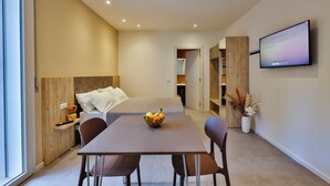 A modern hotel room with a bed, a dining table with chairs, a fruit bowl, a TV mounted on the wall, and a small plant on the side. - Apho Apartment-Hotel (Rimini)