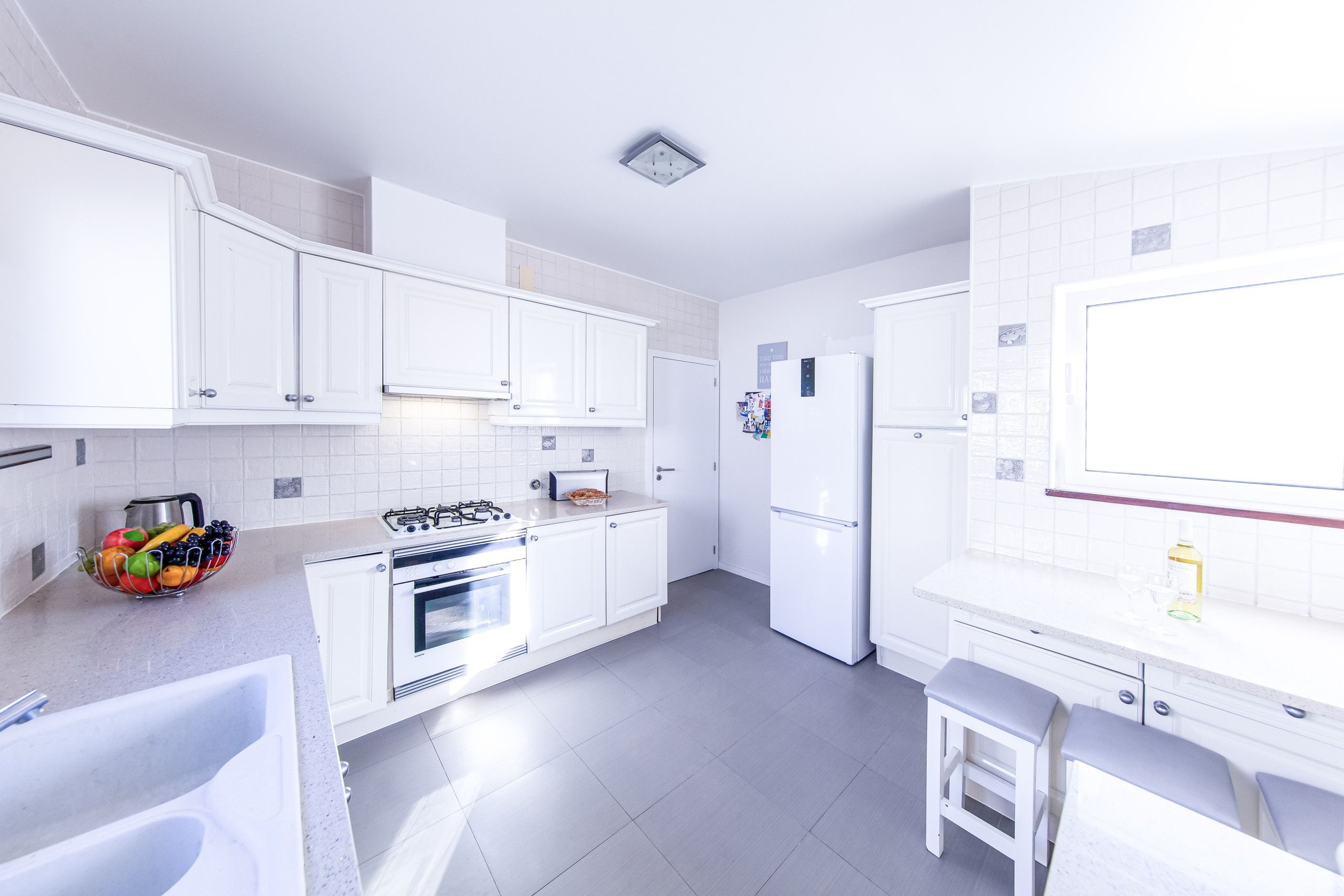 A modern kitchen with white cabinets, a gas stove, an oven, a refrigerator, and a small dining area with a stool.