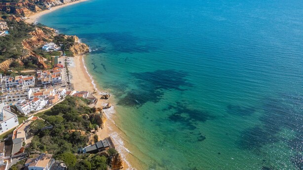Egy tengerparti terület légi felvétele, amelyen egy tiszta, homokos strand, a part mentén elhelyezkedő épületek és a távolban egy hegyvonulat látható.