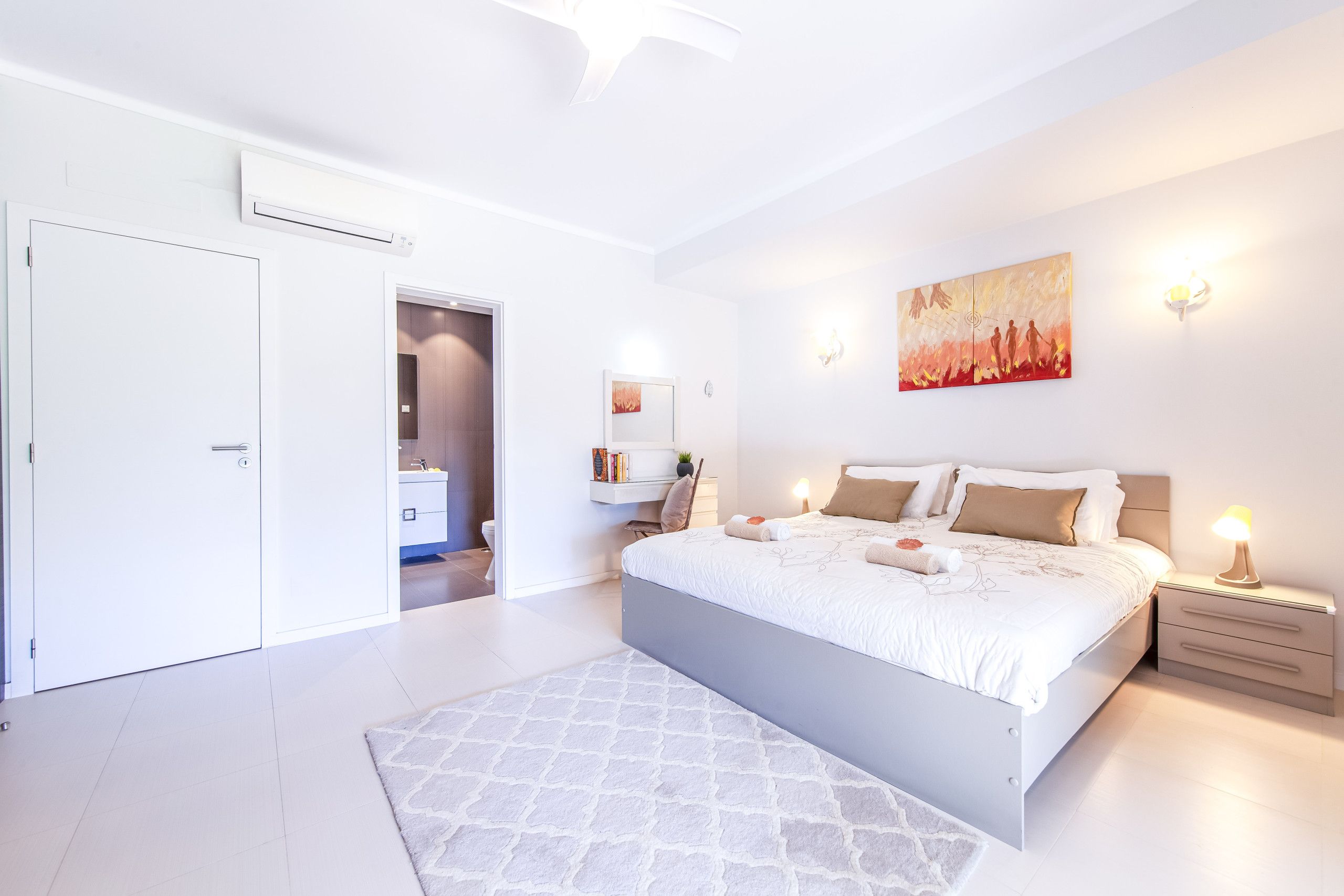Un dormitorio moderno con una cama grande, mesitas de noche, un archivador y una obra de arte en la pared, además de una puerta que da a un baño.