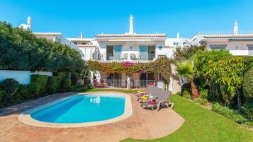 Zona de piscina con suelo de baldosas, rodeada de abundante vegetación y un edificio blanco con varios balcones.