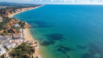 An aerial view of a coastal area with a clear beach, buildings along the shore, and a distant mountain range.