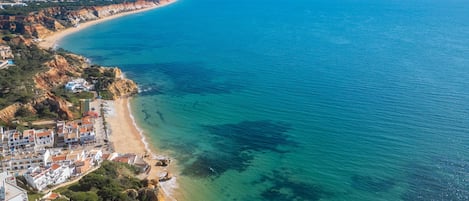 An aerial view of a coastal area with a clear beach, buildings along the shore, and a distant mountain range.