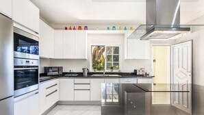 Cocina moderna con armarios blancos, electrodomésticos de acero inoxidable, encimera de vidrio y ventana con vistas a zonas verdes.