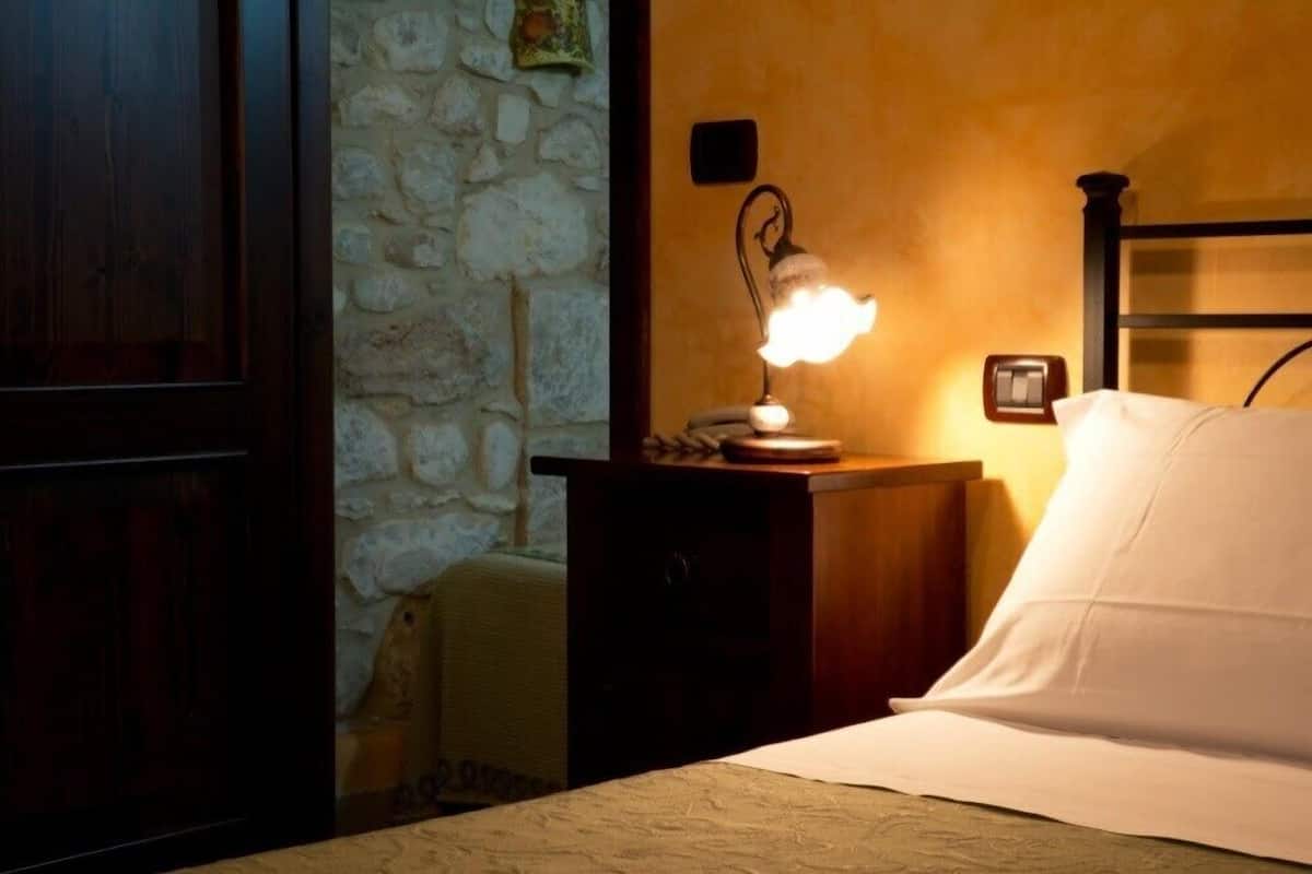 A bedroom with a wooden headboard, a bedside lamp, a stone wall, and a bed with white bedding.