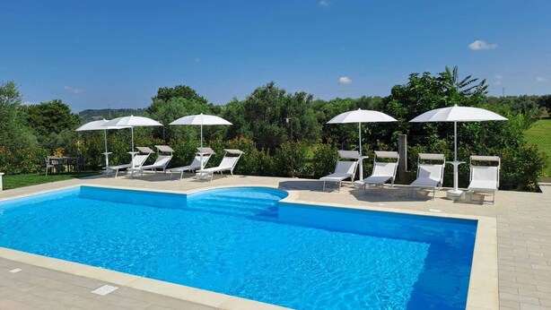 A rectangular swimming pool with clear blue water, surrounded by white lounge chairs and umbrellas. - Agriturismo Il Casolare Di Nonno Mario  (Manoppello)
