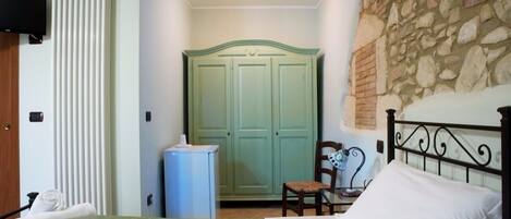 Ein Schlafzimmer mit einem Bett, einem Sessel, einem Schreibtisch, einem Heizkörper, einem Kleiderschrank und einer Steinwand.