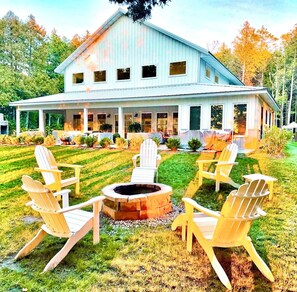 Ein großes Haus mit überdachter Veranda, Feuerstelle und mehreren Adirondack-Stühlen.