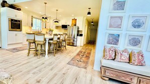 Una cocina moderna con armarios blancos, un comedor con sillas de madera y un pasillo que conduce a una sala de estar con sofá y cojines decorativos.