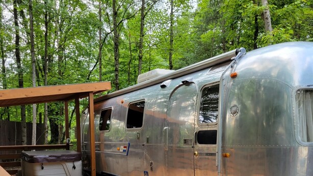 Un camping-car argenté est garé dans une zone boisée, avec une terrasse en bois et un jacuzzi.