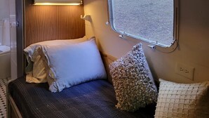 Un intérieur de camping-car compact comprenant un lit, des oreillers, une étagère avec une photo et une fenêtre donnant sur une forêt.