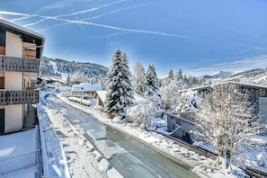 A snow-covered street with buildings on either side, a clear sky, and a large evergreen tree in the center.