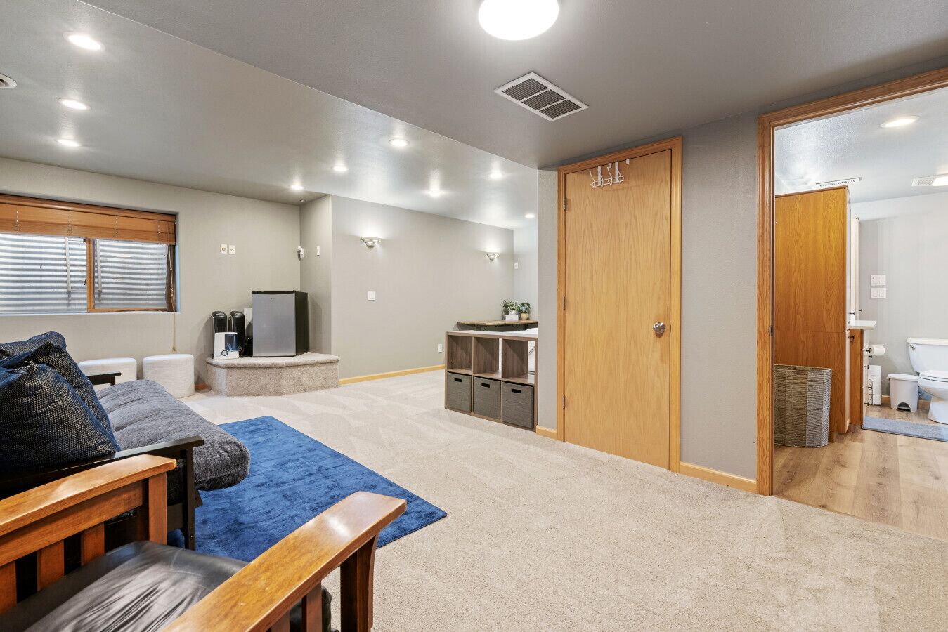 Amplia sala de estar con sofá, una mesa de centro y una alfombra azul. Hay una puerta de madera que da a un baño, y una pequeña zona de kitchenette con refrigerador y microondas.