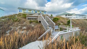 A coastal house with a wooden staircase leading to a deck, surrounded by tall grasses and a cloudy sky. - Charming 5-bedroom oceanfront duplex with wonderful views (Emerald Isle)