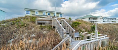 A coastal house with a wooden staircase leading to a deck, surrounded by tall grasses and a cloudy sky.