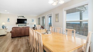 A spacious living room with a wooden dining table, comfortable seating, a flat-screen TV, and a large window with blinds. - Charming 5-bedroom oceanfront duplex with wonderful views (Emerald Isle)