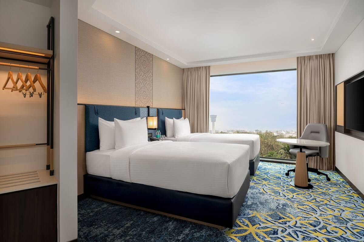 Habitación de hotel con dos camas, un escritorio, una silla, un televisor y un ventanal grande con vista a la ciudad.