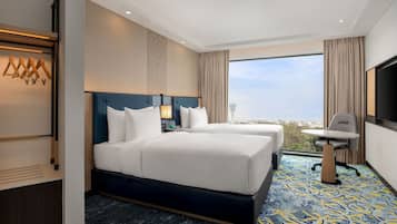 Habitación de hotel con dos camas, un escritorio, una silla, un televisor y un ventanal grande con vista a la ciudad.