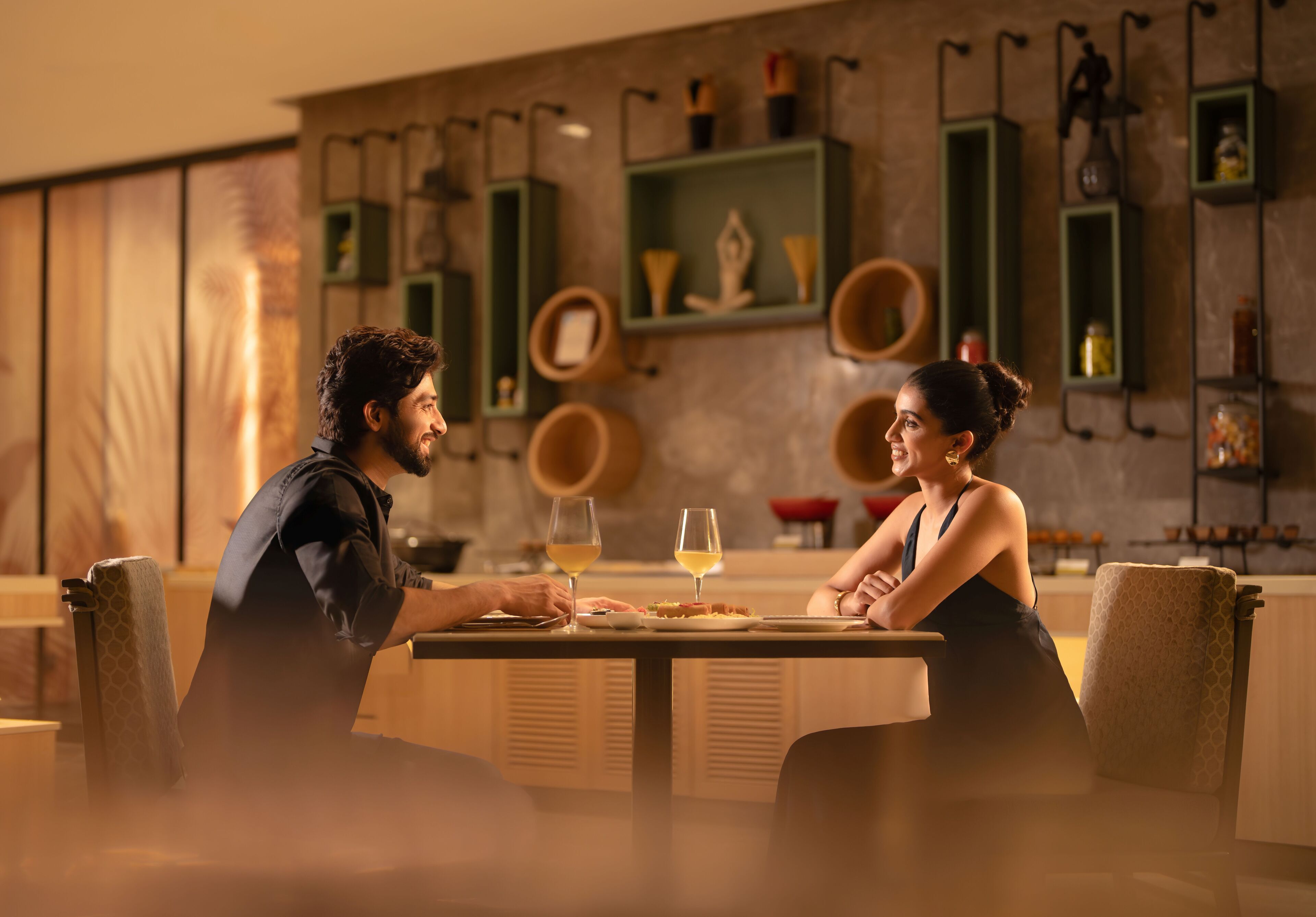 A couple dining at a table in a modern restaurant with a warm ambiance, wooden accents, and green shelving units in the background.