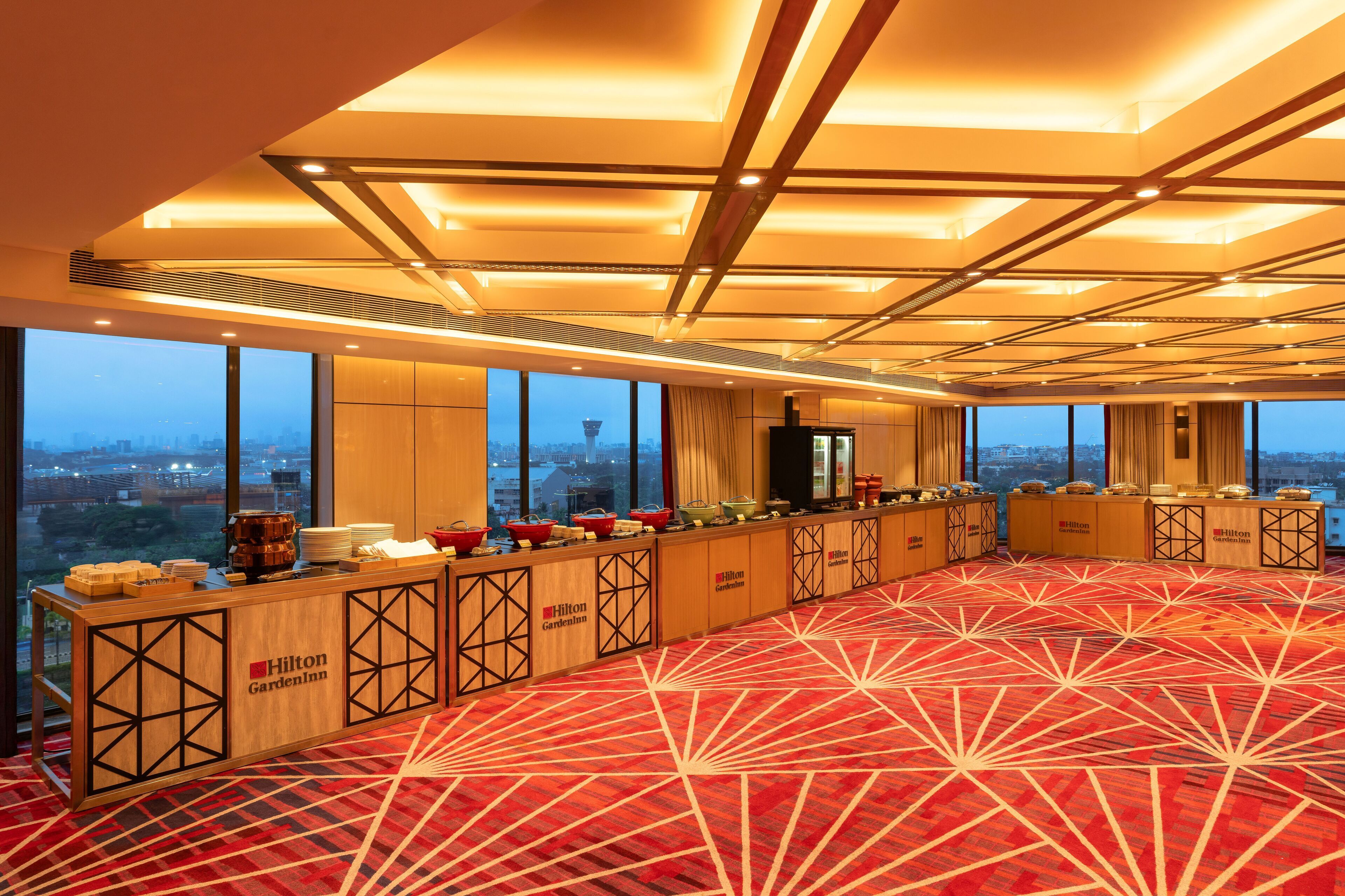 A spacious room with a red patterned carpet, a long buffet table with food items, and large windows offering a city view.