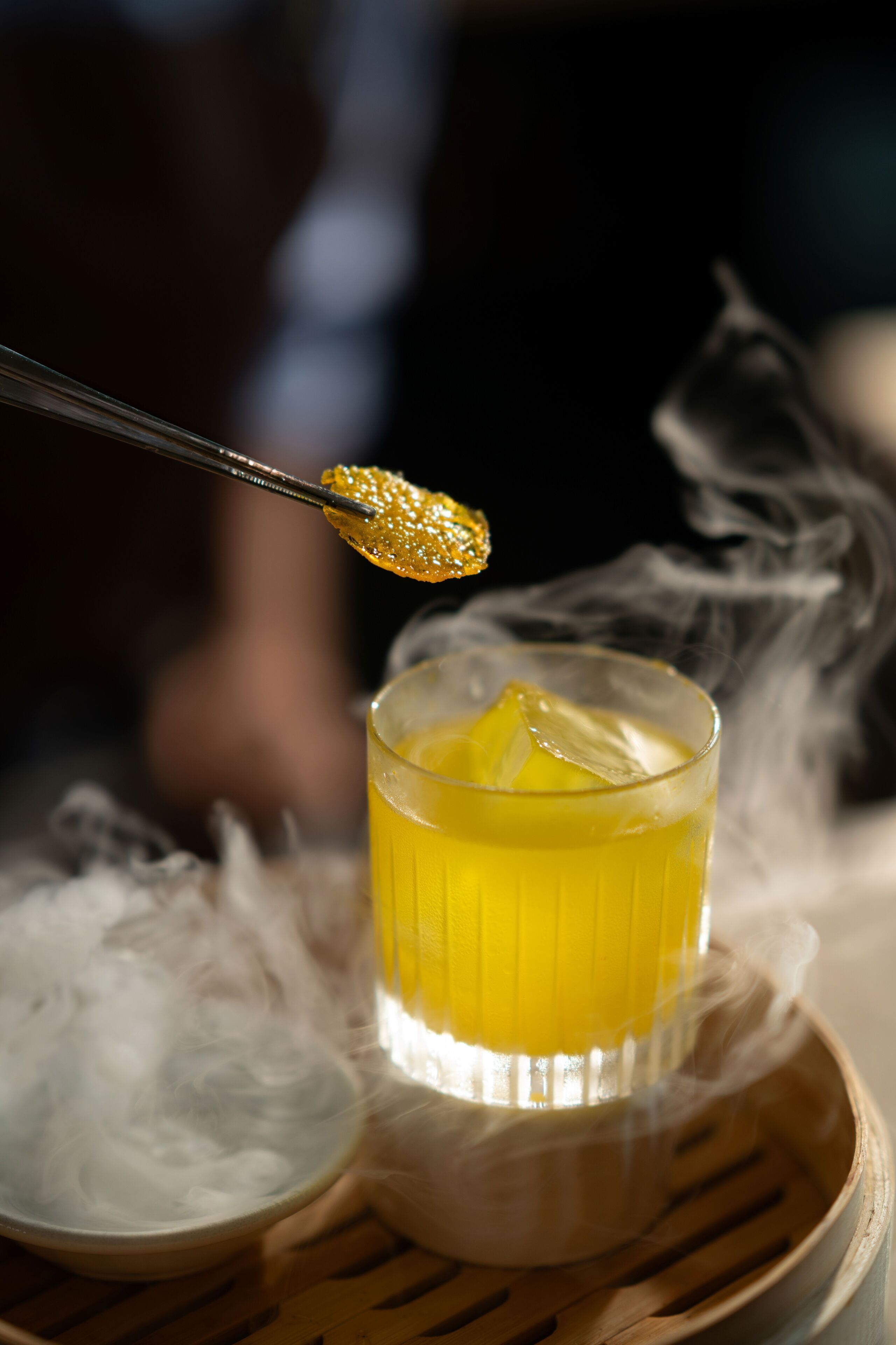 A glass of yellow liquid with ice cubes, a spoon with a golden object, and smoke in the background.