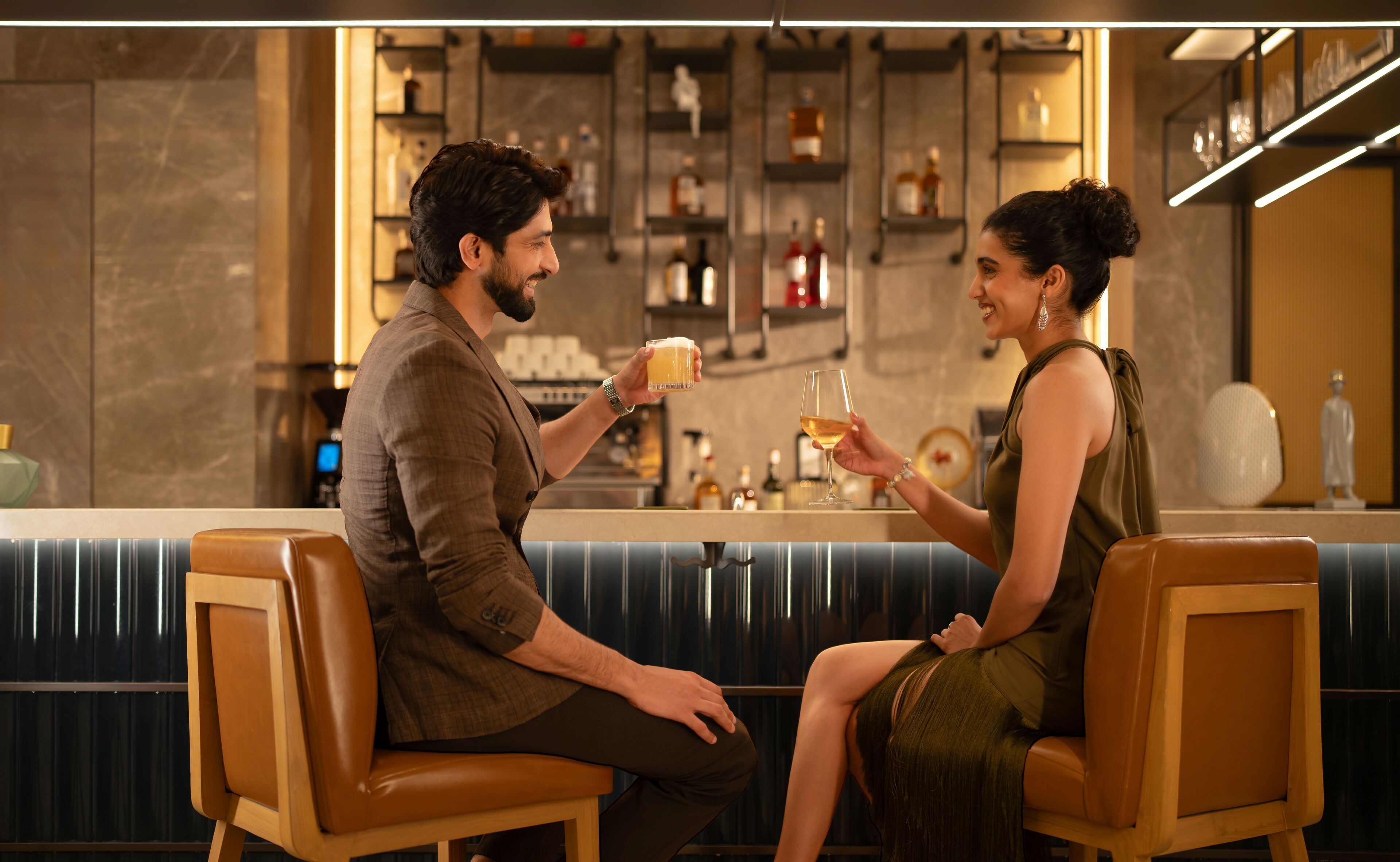 A couple seated at a bar with a modern interior, including a counter with various bottles and a decorative statue.