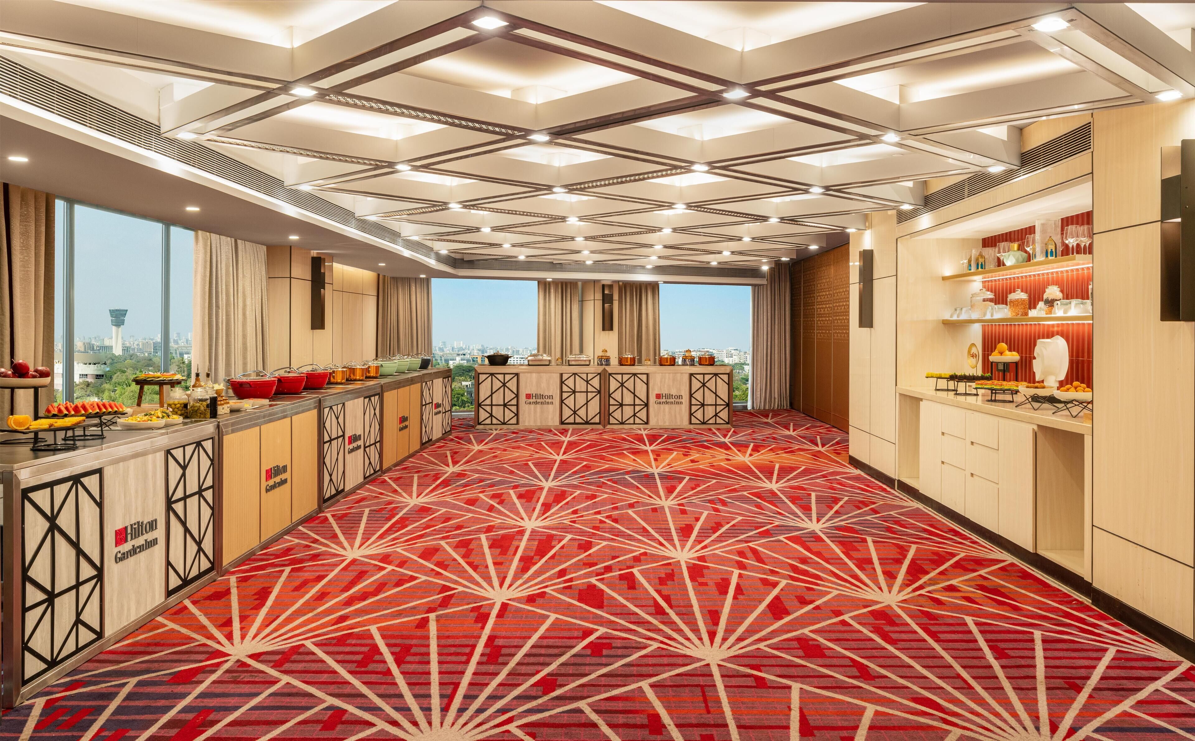 A hotel room with a red patterned carpet, a minibar, and a view of the cityscape.
