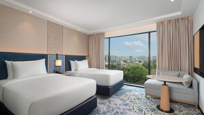 Quarto de hotel com duas camas, um sofá, uma mesa e uma grande janela com vista para a cidade.