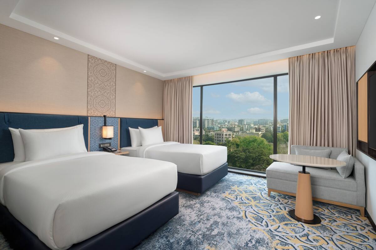 Habitación de hotel con dos camas, un sofá, una mesa y un ventanal con vista a la ciudad.