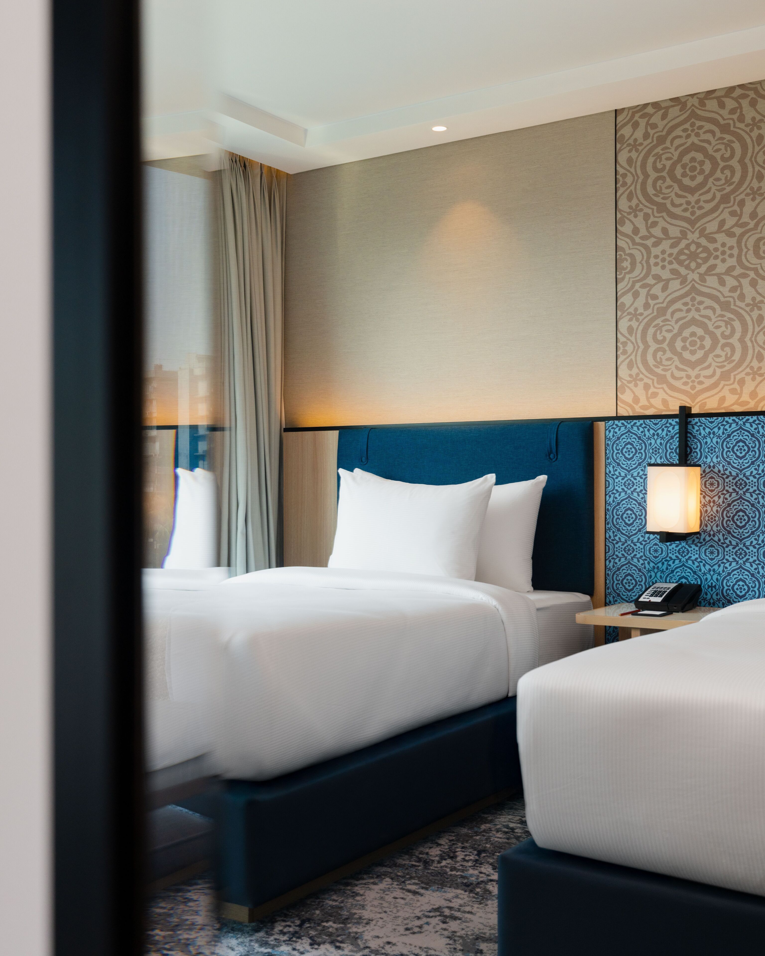 A hotel room with two beds, a nightstand with a lamp, and a patterned wall.